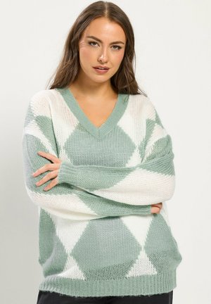 Jeune femme aux longs cheveux bruns portant un pull en maille ample avec un motif de losanges verts et blancs, debout les bras croisés.