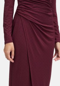 Gros plan sur des mains et une robe bordeaux à manches longues, décorée de petites perles noires et d'un détail de tissu drapé au niveau de la taille.