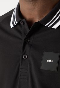Gros plan d'un polo noir avec un col rayé blanc, des boutons noirs et un patch carré noir avec le mot BOSS en lettres blanches.