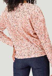 Haut à sequins roses avec manches longues, présentant une coupe ample et une texture chatoyante. Porté par-dessus un pantalon en denim foncé.