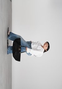 Moteris, vilkinti baltą palaidinę ir mėlynus džinsus, laiko didelę juodą rankinę, stovėdama prieš paprastą šviesiai pilką foną.