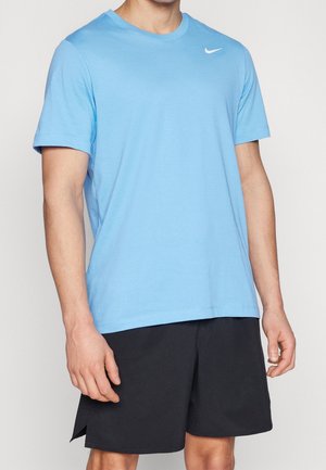 Mężczyzna ubrany w jasnoniebieską koszulkę Nike z krótkim rękawem i czarne sportowe szorty, stojący na tle jednolitego jasnego tła.