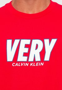 Raudonas bliuzonas su dideliu baltu užrašu "VERY", apibrėžtu mėlyna spalva, ir mažesniu baltu užrašu "Calvin Klein" po juo krūtinės srityje.