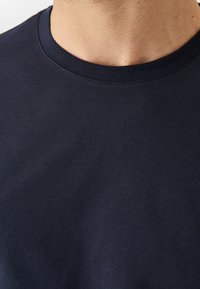 Primo piano del collo e della parte superiore del torace di una persona che indossa una maglietta a maniche corte a girocollo di un blu scuro uniforme.