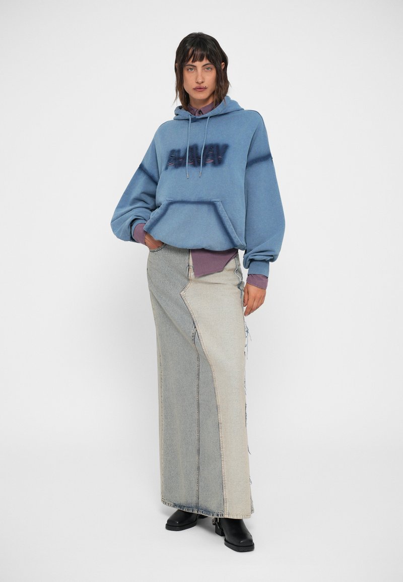 Blå hættebluse med slidt skrift, lagd over en lilla skjorte, parret med en lang, asymmetrisk lys denimskørt. Sorte ankelstøvler.