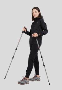 Veste imperméable noire avec capuche, associée à un pantalon noir et des chaussures de randonnée grises. L'utilisateur tient deux bâtons de randonnée réglables.