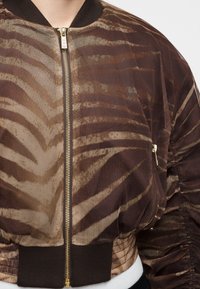 Κοντινό πλάνο καφέ διαφανούς μπουφάν bomber με σκούρες ρίγες ζέβρας, χρυστό φερμουάρ και μαύρο ριμπ γιακά και τελείωμα.