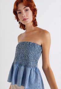 Denim stroppeløs topp med smockert liv og flagrende peplum nederkant. Har lyseblå stoff med tekstur og bølget kant.