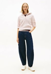 Femme debout avec la main dans la poche, portant un pull en tricot rose clair, un jean large noir et des baskets blanches, sur fond blanc.