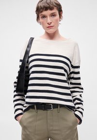 Οριζόντια ριγέ πουλόβερ σε κρέμα και ναυτικό, χαλαρή εφαρμογή, με χαμηλούς ώμους. Συνδυασμένο με ανοιχτό καφέ παντελόνι και μαύρη τσάντα.