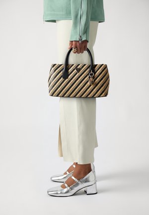 Person wearing cream pants and silver shoes holds a black and beige striped handbag with black handles and gold charms.