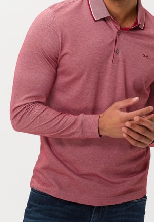 Mann trägt ein rotes Poloshirt mit langen Ärmeln und Knöpfen, verschränkt die Hände und trägt dunkelblaue Jeans vor einem einfarbigen Hintergrund.