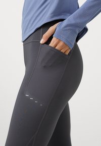 Hoge taille, donkergrijze legging met een zijzak, een zachte textuur en reflecterende details langs de onderbenen, gecombineerd met een lichtblauwe longsleeve top.