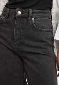 Jeans de denim negro de cintura alta, con un cierre de botón plateado, dos bolsillos frontales y una textura ligeramente desgastada.