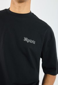 Black cotton t-shirt with short sleeves and a crew neck, featuring white Gothic script "Mystoria" on the left chest area.