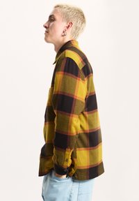 Camicia in flanella con motivo a quadri giallo, nero e rosso. Presenta un colletto classico, maniche lunghe e polsini con bottoni. Texture morbida visibile.