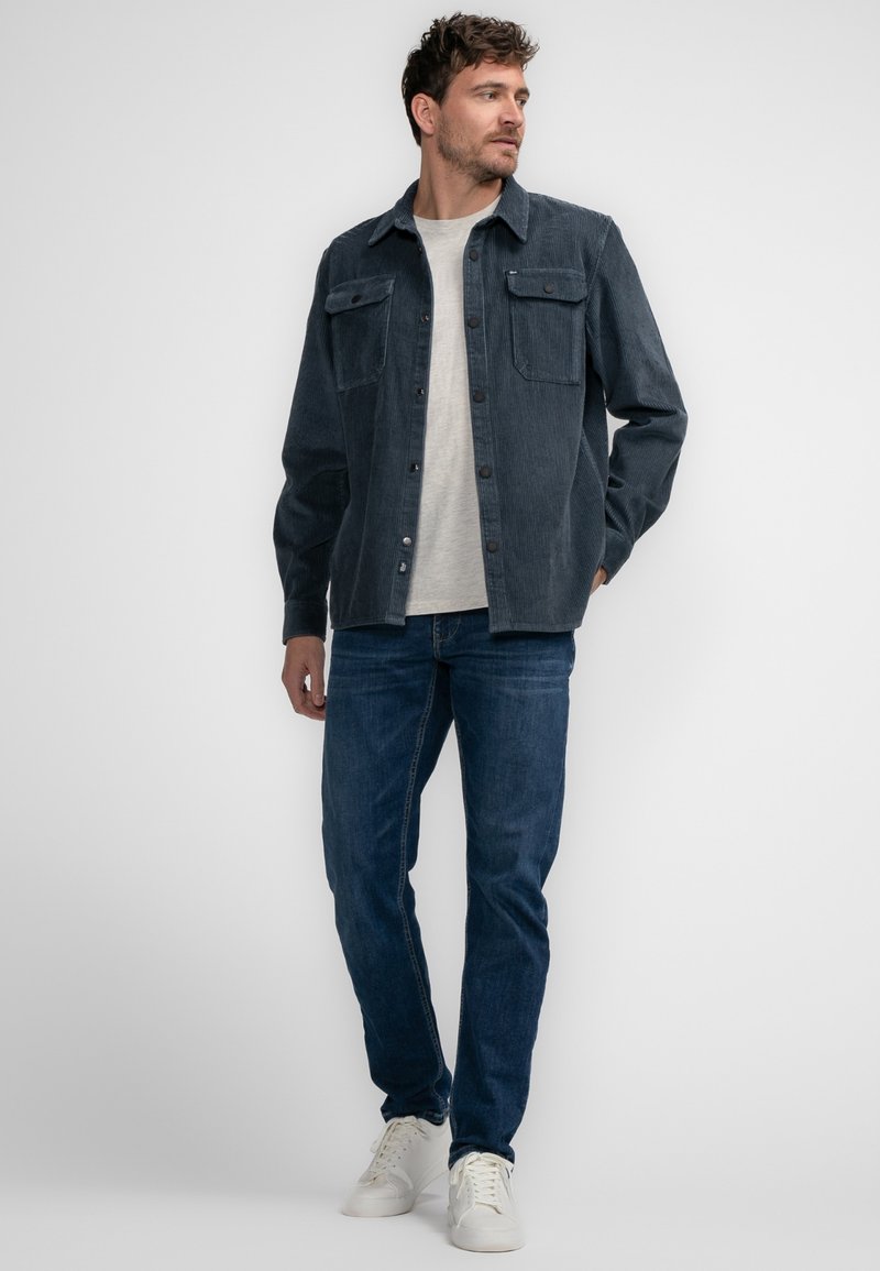 Veste en velours côtelé bleu marine avec deux poches poitrine, fermeture par boutons, manches longues, associée à un jean bleu et des baskets blanches.