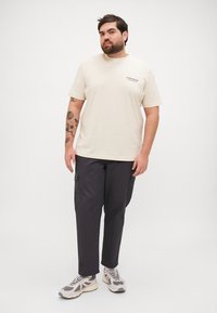 Beige bomull T-skjorte med korte ermer og trykt tekst; parret med mørke cargo-bukser med sidelommer; grå og hvite joggesko.