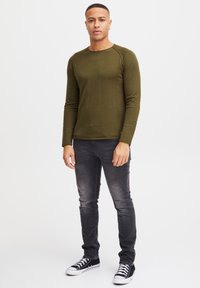 Olijfgroene longsleeve met ronde hals, gecombineerd met donkere slim-fit jeans en zwarte sneakers met witte accenten.