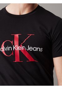 Svart t-shirt med stora röda "CK"-bokstäver och vit text "Calvin Klein Jeans" över bröstet, bärs av en person med ljus hud.