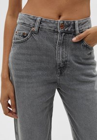 Los jeans de mezclilla gris presentan una cintura alta, un cierre de botón de cobre y un diseño clásico de cinco bolsillos, con una textura suave y ligeramente desgastada.