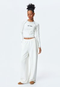 Weißes, geripptes Langarmoberteil mit dem Text "NY 1982", kombiniert mit lockeren weißen Hosen. Der Stoff wirkt weich; das Design umfasst kontrastierende Nähte.