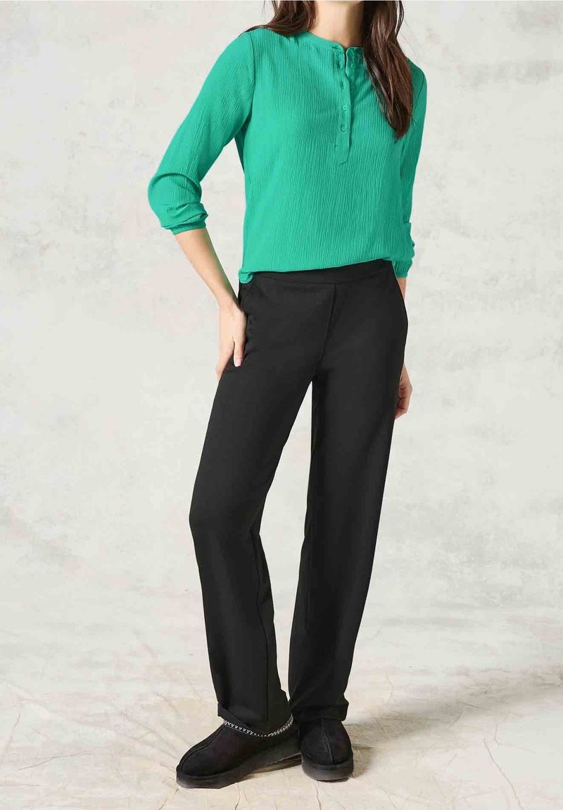 Femme portant un chemisier vert boutonné à manches trois-quarts, un pantalon noir droit et des chaussures noires sans lacets, debout avec une main dans la poche.