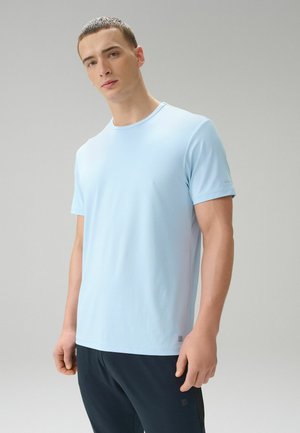 Ung mand med kort hår iført en lyseblå kortærmet skjorte og mørke bukser, stående foran en ensfarvet grå baggrund.