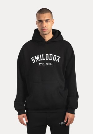 Svart huvtröja i mjukt material med en framficka, försedd med vit text "SMILODOX ATHL. WEAR" över bröstet och ribbade muddarna.