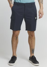 Mørkeblå cargo shorts laget av bomull, med front- og side lommer, knappelukking og en vevd etikett på siden. Teksturert stoff.