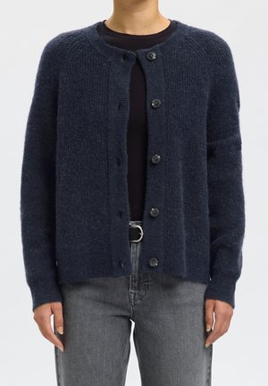 Personne portant un cardigan bleu foncé boutonné par-dessus un haut noir, associé à un jean gris et une ceinture noire avec une boucle argentée.