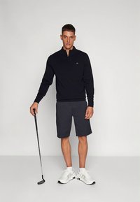 Tengerészkék cipzáros pulóver, szürke rövidnadrág és fehér sportcipő. A modell golfütőt tart, semleges szürke háttér előtt áll.