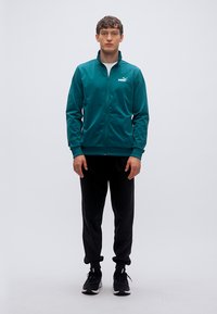 Τζάκετ αθλητικό με φερμουάρ σε τιρκουάζ χρώμα και μακριά μανίκια, μαύρο αθλητικό παντελόνι και μαύρα αθλητικά παπούτσια. Μαλακό ύφασμα με λεία υφή. Λογότυπο Puma στο στήθος.