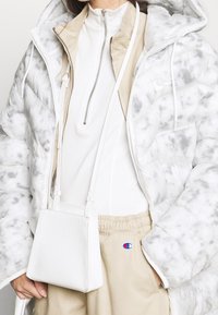 Uppblåst vit jacka med grå mönster, beige zip-up inre lager och ljusbeige byxor. Liten vit texturerad väska med en lång rem.