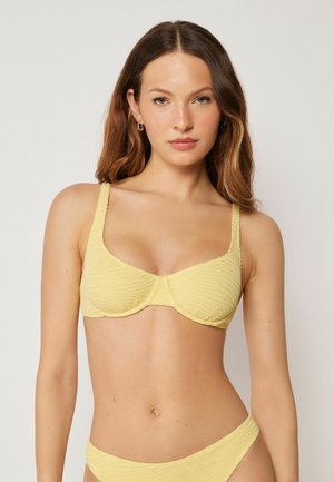 Kvinde med langt brunt hår iført en tekstureret gul bikini top og bund, stående mod en ensfarvet lys baggrund.