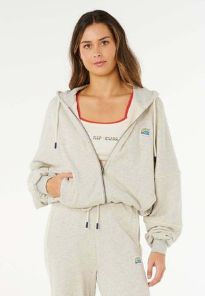 Frau trägt hellgrauen Kapuzenpullover mit Reißverschluss und passende Jogginghose mit "Rip Curl"-Logo, steht vor einem schlichten weißen Hintergrund.