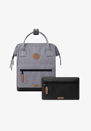 Grauer Stoff-Rucksack mit Frontreißverschlusstasche und schwarzer Leder-Setztasche, beide mit braunen Markenlabels und Zugschnüren.