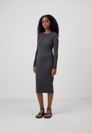 Robe grise à manches longues, ajustée, en matière côtelée, atteignant le mollet. Portée avec des sandales noires plates, dotées de bretelles réglables.