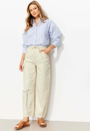 Femme portant une chemise à rayures bleu clair boutonnée, un pantalon large crème avec un motif floral subtil, et des sandales ouvertes couleur sable, debout contre un mur blanc.