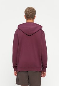 Ung mand med kort krøllet hår står med ryggen til, iført en vinrød hættetrøje og mørkebrune shorts foran en neutral lys baggrund.