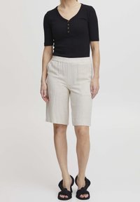 Černý žebrovaný top s krátkým rukávem a knoflíkovou légou, spárovaný s lehkými béžovými kraťasy ke kolenům. Obuv má černý uzlík jako detail.