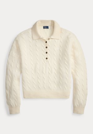 Kvinde står med hænderne i lommerne iført en kremfarvet kabelstrikket sweater, blå jeans, brun bælte og sennepsfarvede ruskindsmokkasiner mod en neutral baggrund.