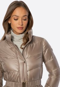 Mujer con cabello largo y castaño, que lleva una chaqueta acolchada beige con cuello alto y un suéter de cuello alto gris claro debajo.