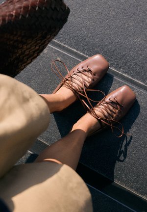 Scarpe stringate in pelle marrone con punte quadrate indossate a piedi nudi, su pavimento piastrellato scuro e abbigliamento beige visibile ai bordi.