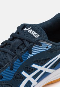 Tummansininen urheilukenkä, jossa on verkkokangasta, valkoisia ja sinisiä vinoviivoja, nauhat ja hopea logo kielen päällä.