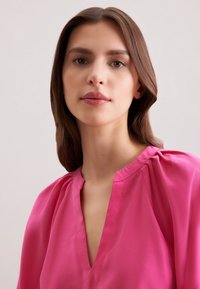 Blusa rosa brillante con scollo a V, tessuto morbido con leggero lucido e dettagli arricciati sulle spalle per aggiungere texture.