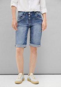 Shorts en denim bleu clair avec un délavage, ourlets retroussés, deux poches avant et double bouton à la taille. Portés avec des baskets.