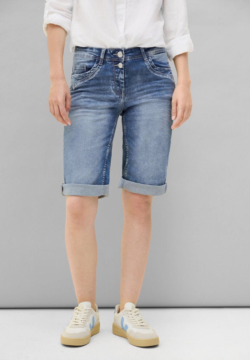 Shorts en denim bleu clair avec un délavage, ourlets retroussés, deux poches avant et double bouton à la taille. Portés avec des baskets.