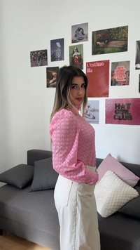 Νέος άνθρωπος με ροζ μπλούζα με σχέδια και λευκό παντελόνι στέκεται πλαγίως σε ένα δωμάτιο με γκρι καναπέ και αφίσες τέχνης στον τοίχο πίσω τους.