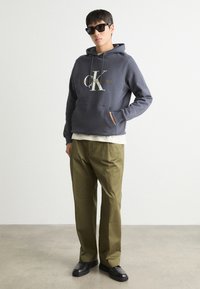 Siva kapuca s puloverjem z logotipom "CK" in besedilom "Calvin Klein Jeans", v kombinaciji z olivno zelenimi hlačami in črnimi čevlji.